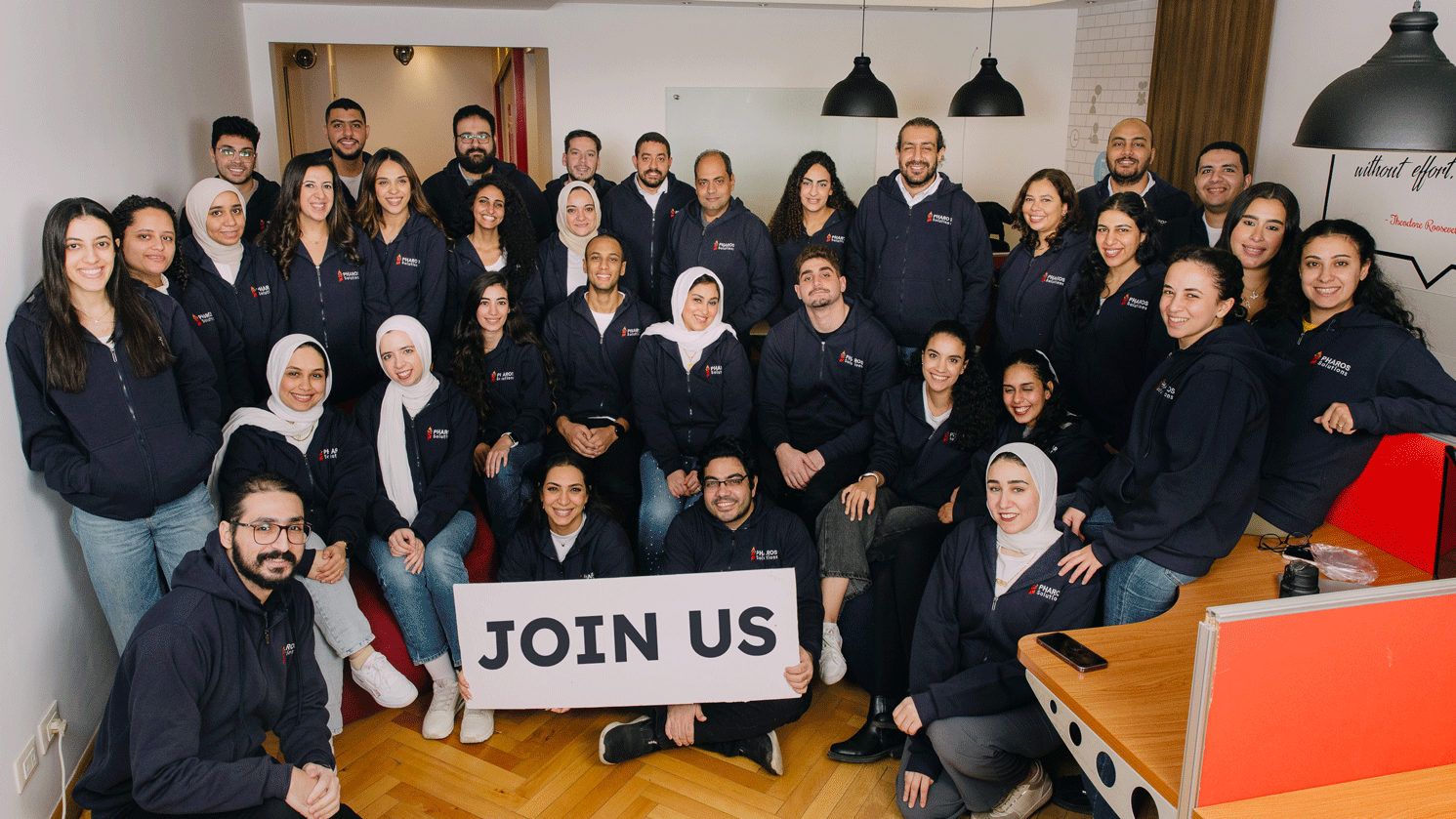 Team of software engineers working at Pharos Solutions holding a sign that says join us to work in a well-established software development company